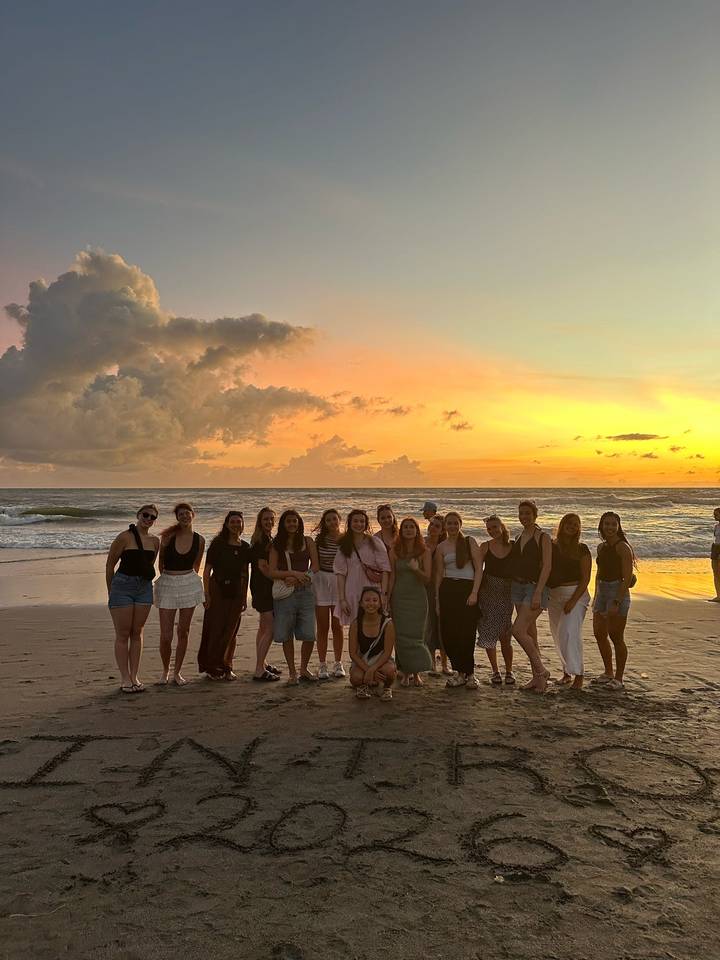 Large group of women posing on a beach during a vivid golden sunset with waves behind.