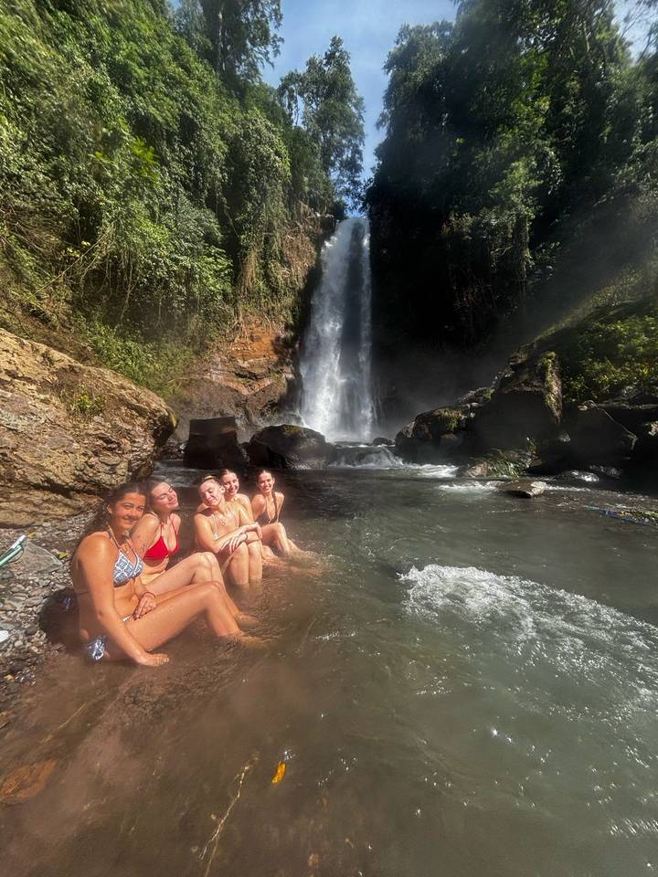 Group of women in swimsuits relaxing in a natural pool at the base of a jungle waterfall.
