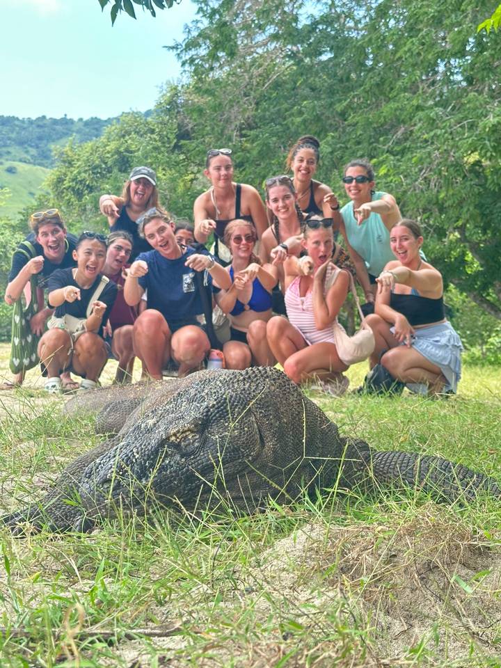 Excited group crouching behind a large Komodo dragon on grassy ground.