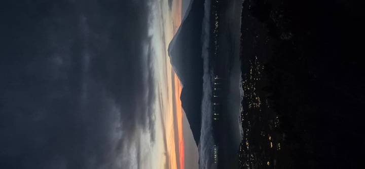 Silhouetted volcano rises above a lake and town lights at dawn with colorful clouds.