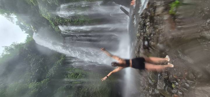 Person with arms raised standing in front of a misty jungle waterfall; image is soft and hazy.