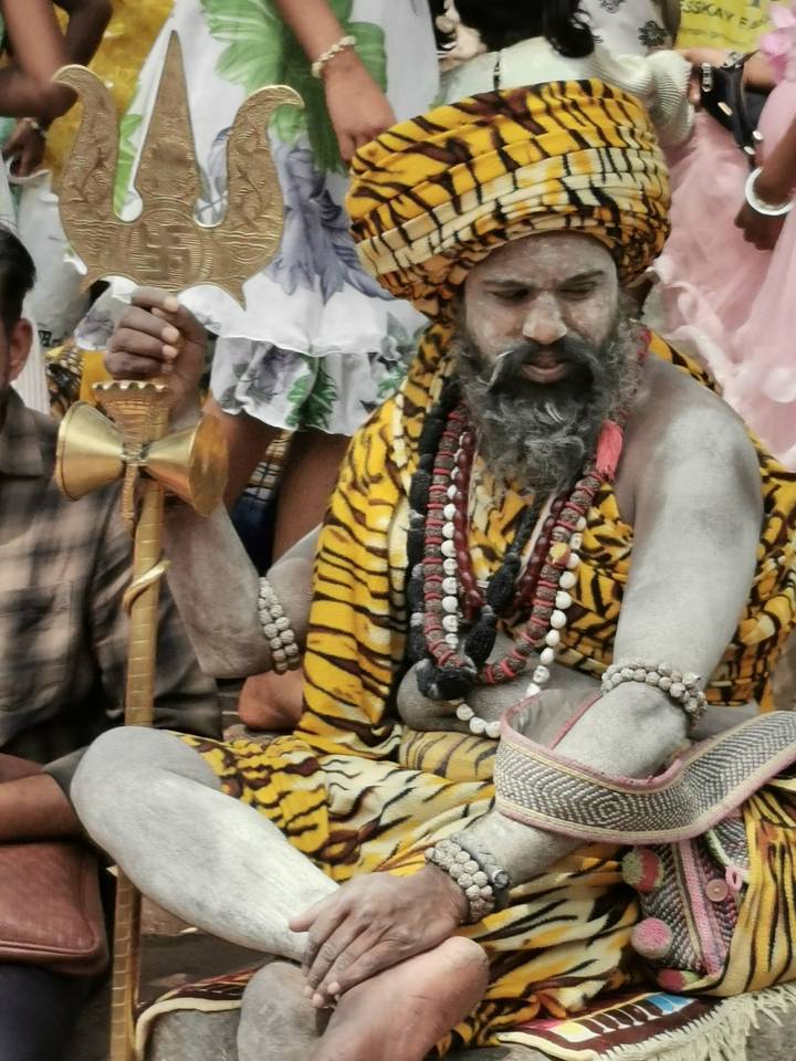 Close-up of a bearded holy man covered in ash, wearing tiger-print cloth and multiple bead necklaces, holding a ceremonial staff.