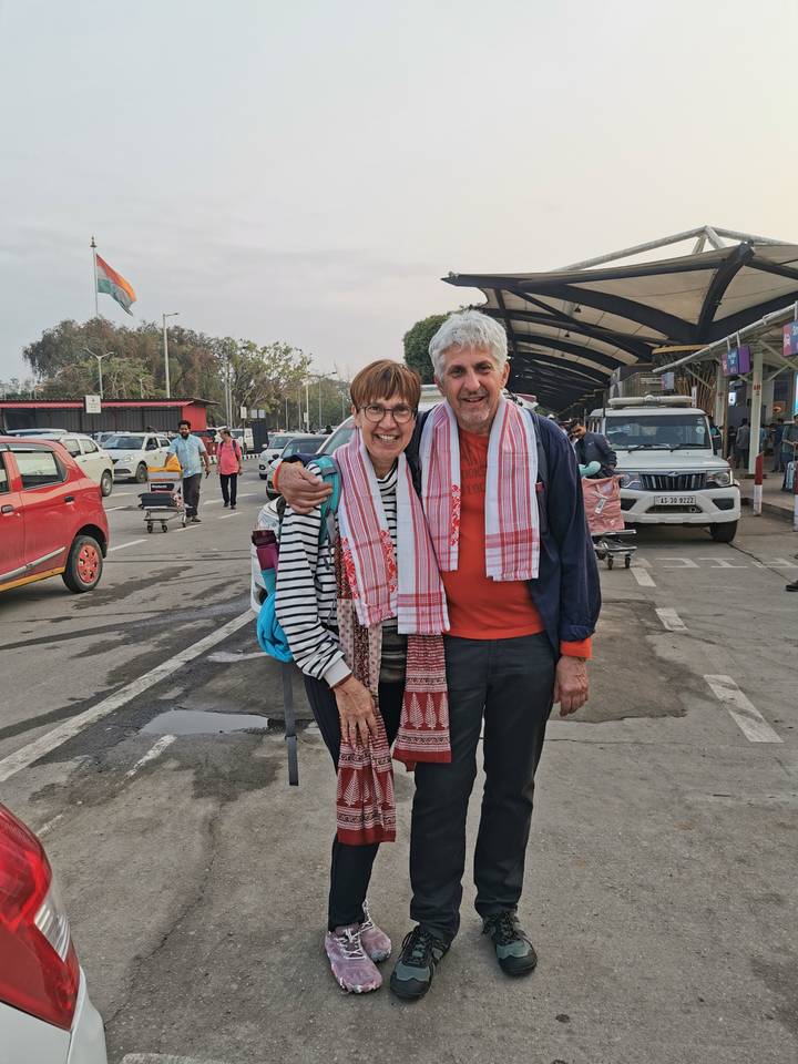 Smiling older couple with Assamese scarves stand arm-in-arm outside an airport terminal with vehicles and trolleys behind them.