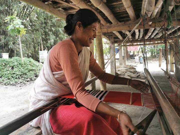 Smiling woman works a traditional handloom beneath a bamboo shelter in a rural setting.