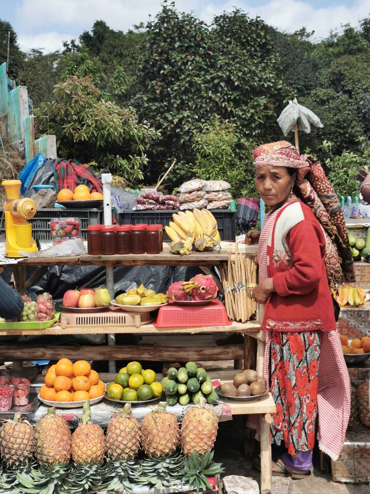 Market vendor in traditional attire stands beside a stall displaying fresh fruit, jarred jam and produce.