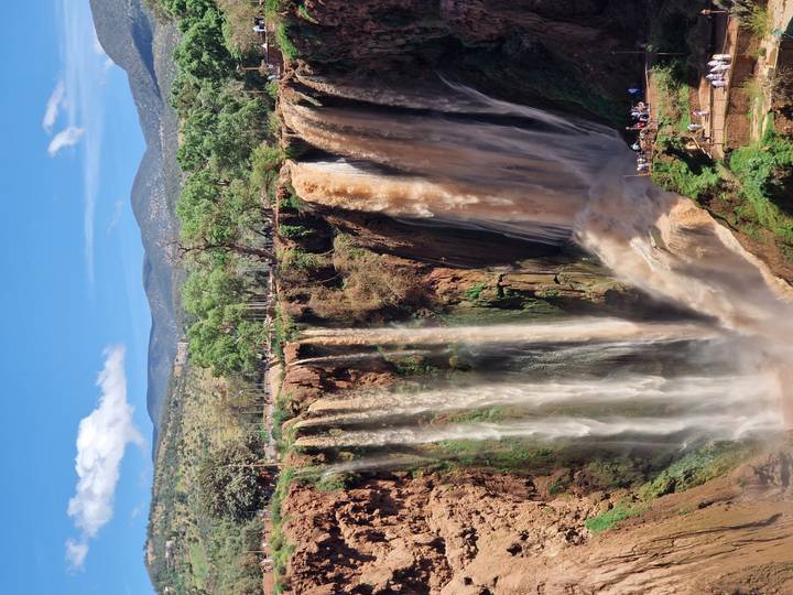 Powerful twin waterfalls plunging over a tall cliff into a lush canyon framed by mountains and blue sky