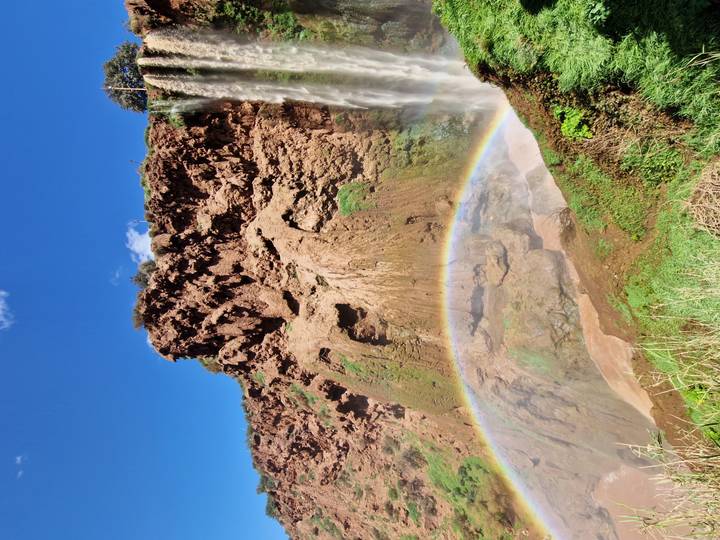 Rainbow arches at the base of an ochre cliff with a waterfall against a vivid blue sky