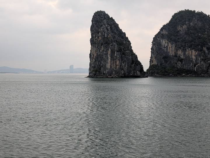 Overcast view of a tall limestone karst rising from calm gray waters with distant city skyline in Halong Bay.