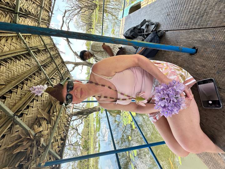 Tourist wearing a floral crown sits on a rustic boat amid lotus-filled wetlands.