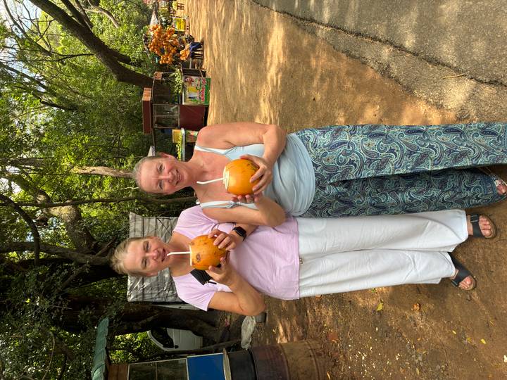 Two travelers smile holding fresh coconuts on a shaded roadside in Sri Lanka.