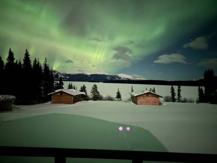 Wide view of frozen lake, cabins and green aurora curtains over mountains