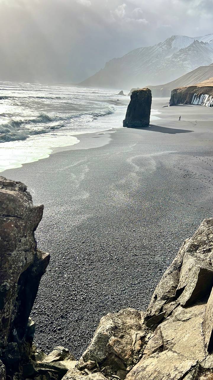 Close-up view of a dark volcanic sand beach with gentle surf and foam lines.