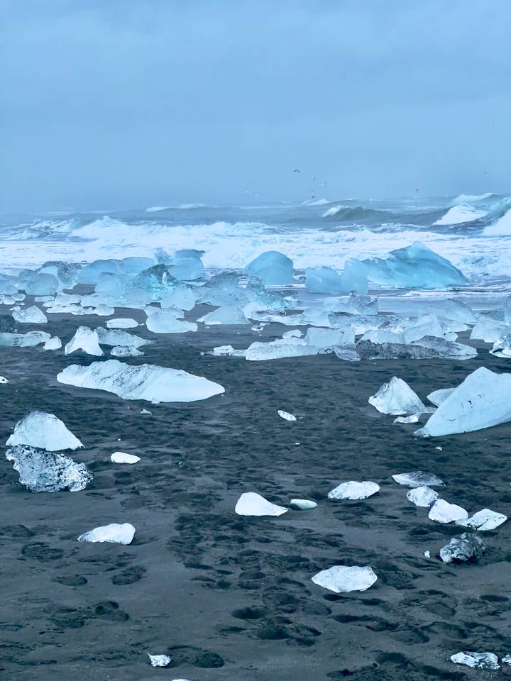 Large blue ice chunks scattered across a black sand shore with waves crashing behind.