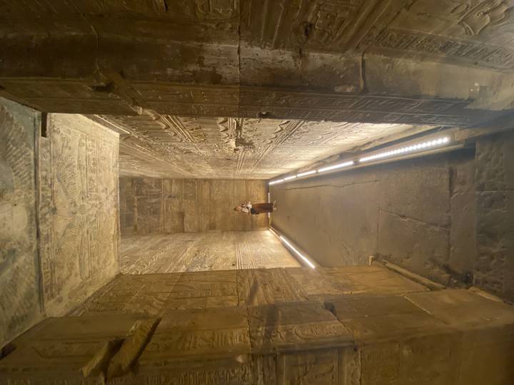 Dimly lit stone corridor with reliefs and a lone visitor in the distance.