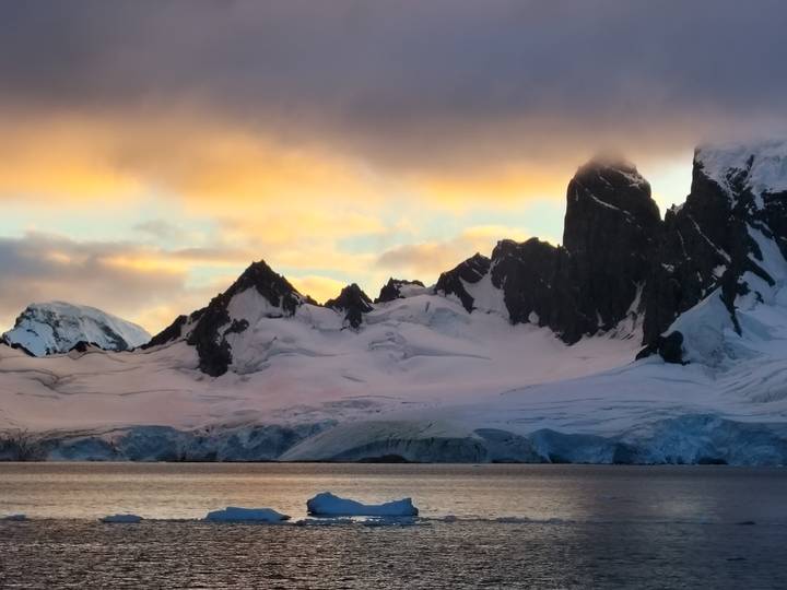 Snow-clad Antarctic peaks at sunset with dramatic clouds glowing orange and pink.