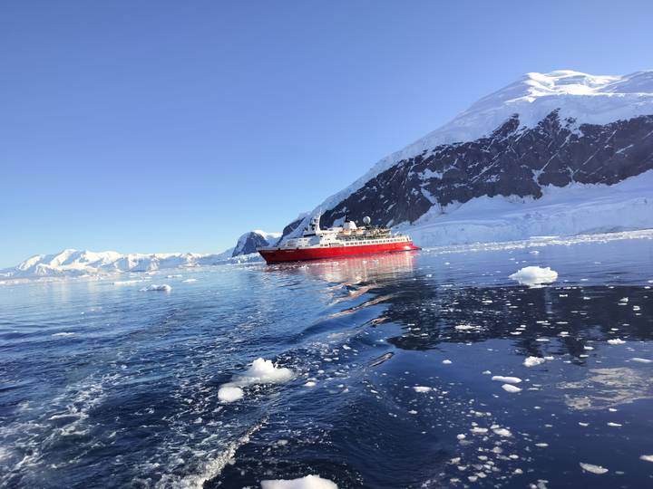 Red expedition ship cruising through icy Antarctic waters beneath towering glacier cliffs.