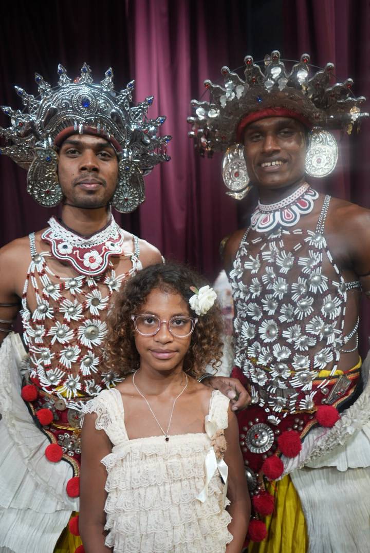 A young girl poses between two traditionally dressed Sri Lankan dancers wearing ornate silver chest pieces.
