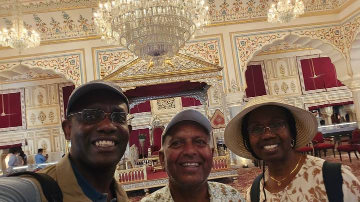 Three smiling travellers posing inside an ornately decorated palace hall with chandeliers.