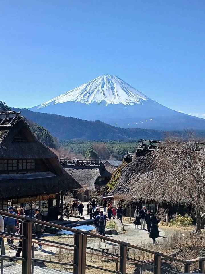Traditional thatched village houses lie beneath the majestic snowy cone of Mount Fuji on a clear day.