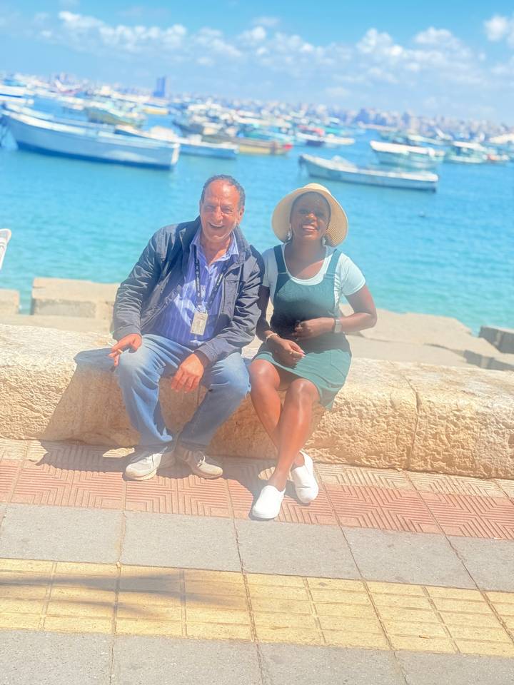 Smiling man and woman sit on seaside stone wall with turquoise water.