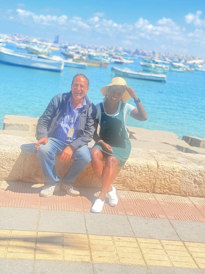 Two travelers pose on stone ledge with bright blue Mediterranean behind.