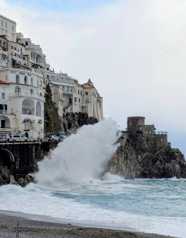 Powerful wave crashes against cliffside buildings of the Amalfi Coast on a cloudy day.