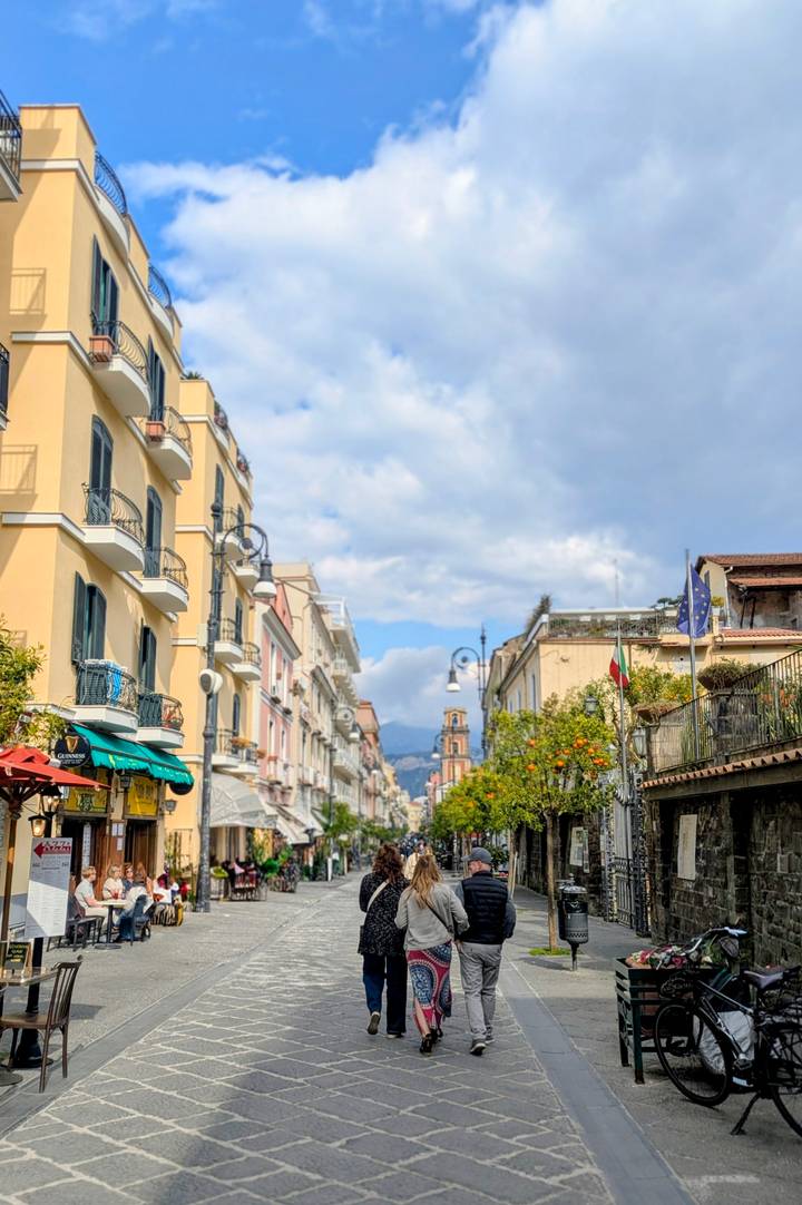 Charming pastel buildings line a pedestrian street in Sorrento with citrus trees and distant mountains.