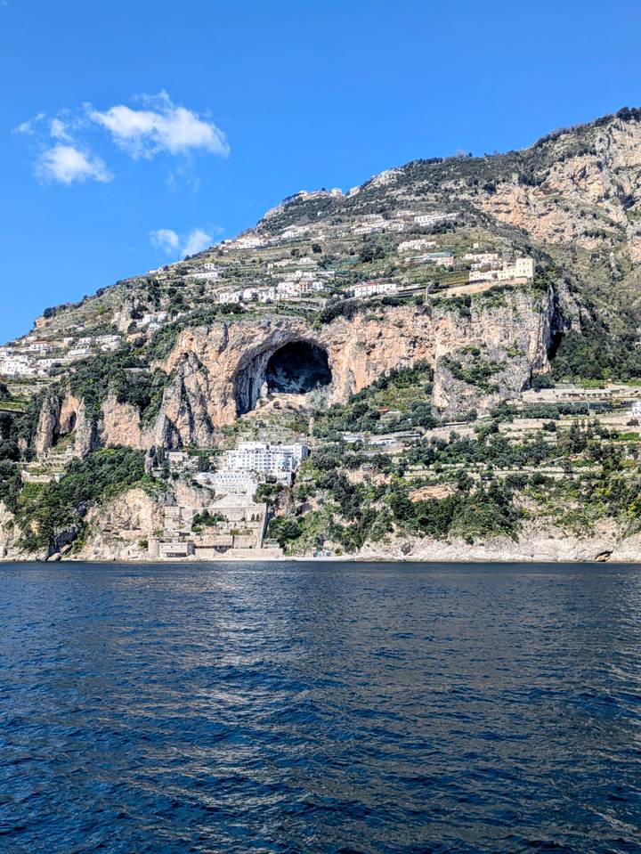 Massive cliff with a vast cave opening rising above terraced slopes along the Amalfi coastline.