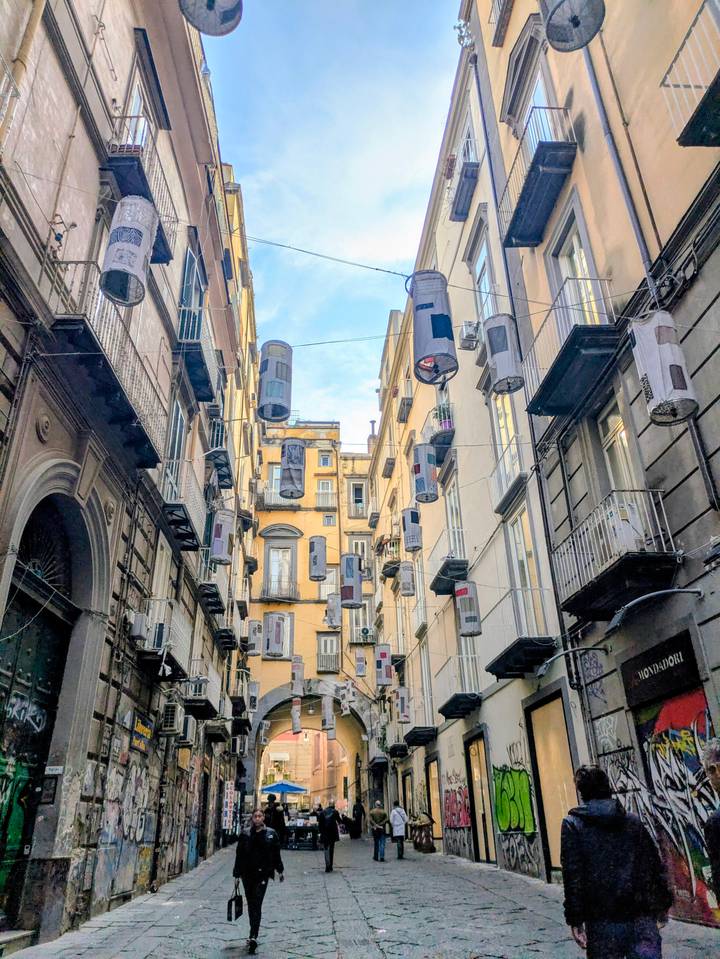 Quirky art installations hang between narrow historic buildings on a lively lane in Naples.