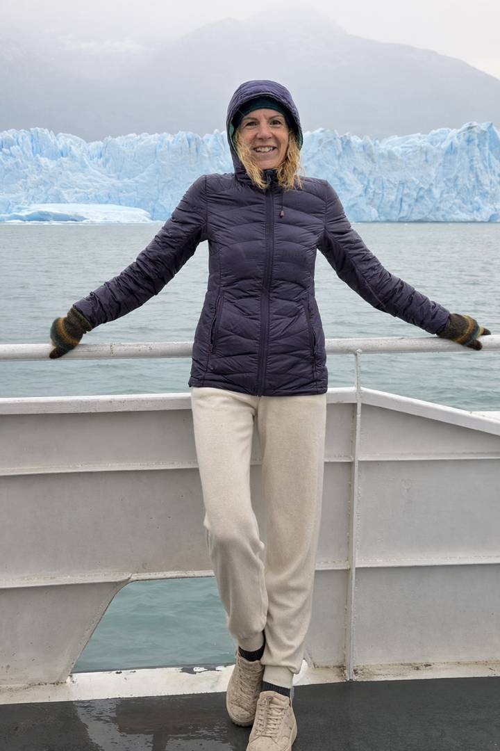 Woman in a puffer jacket poses with arms outstretched on the deck of a boat with cold grey water behind.