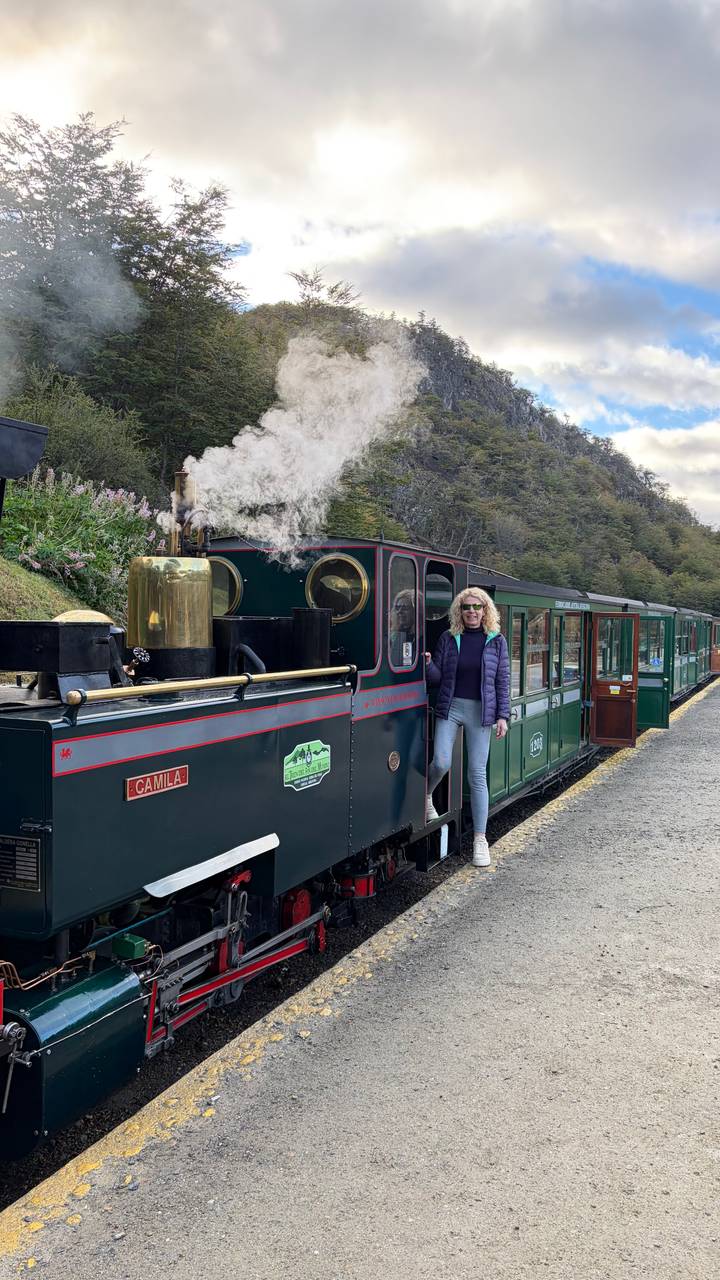 Traveler stands beside a vintage steam train in a forested station with steam billowing into the air.