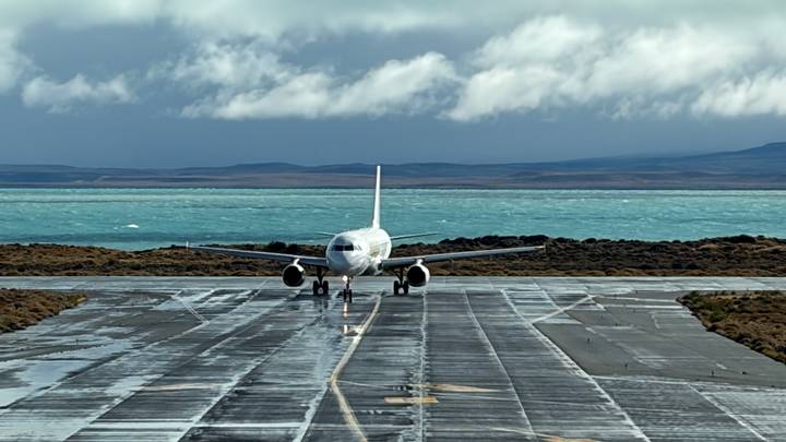 Small passenger jet taxis along a rain-slicked runway beside turquoise lake waters and dramatic skies.