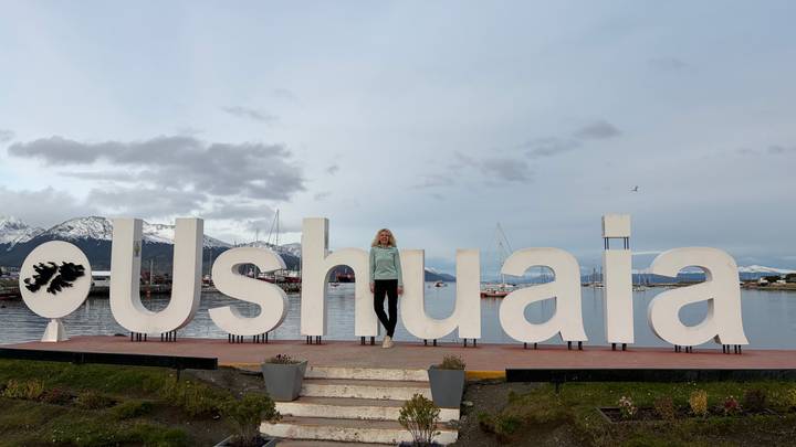 Traveler poses within the giant white Ushuaia sign on the waterfront with snowy peaks beyond.