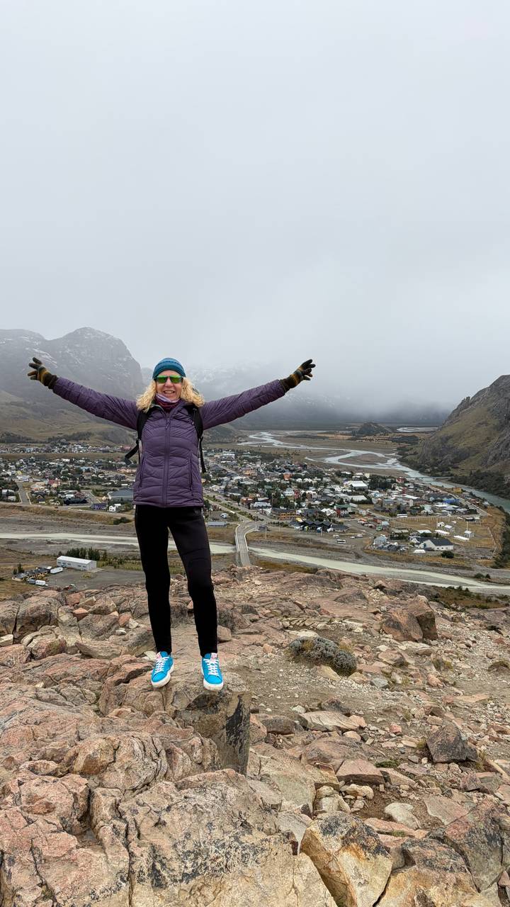 Excited hiker raises arms high above a Patagonian town stretching along a river valley framed by peaks.