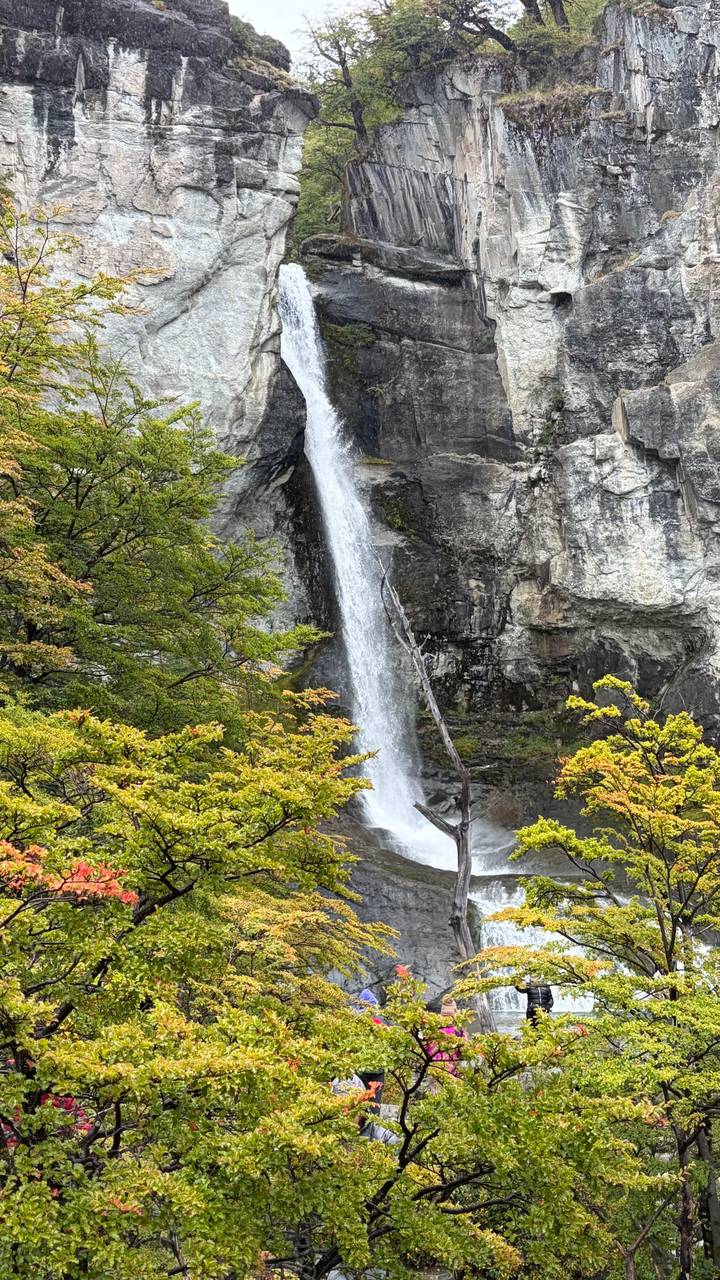 Tall narrow waterfall cascades down a rocky cliff surrounded by early autumn foliage.