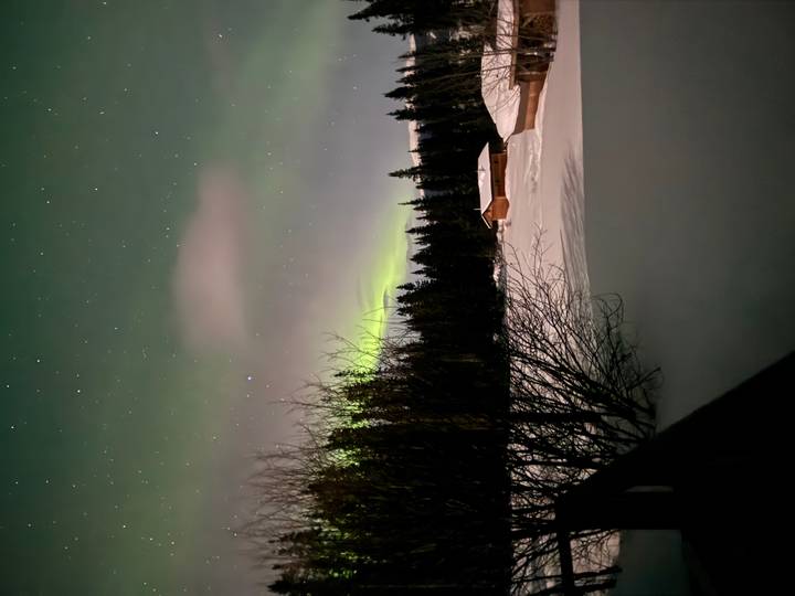 Subtle green aurora glow above snowy ground and a small wooden cabin framed by dark trees.