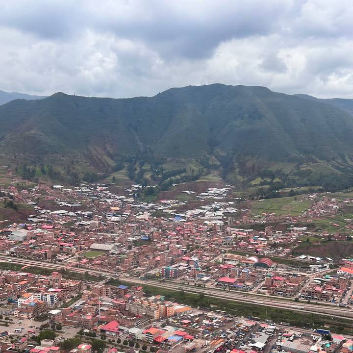A sprawling Andean town of red roofs fills a valley below towering green mountains under a cloudy sky.
