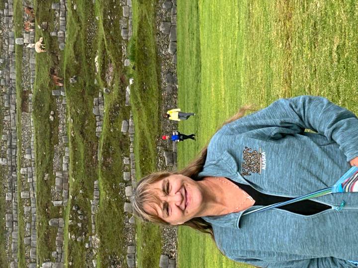 Smiling woman in front of stone agricultural terraces and grassy field with a few llamas grazing in the background.