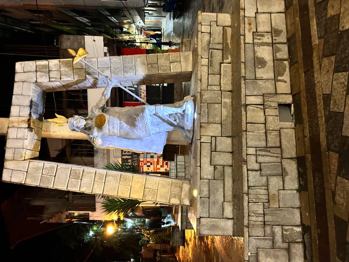 Night-time statue of an Inca ruler holding a staff, illuminated in a small town square.