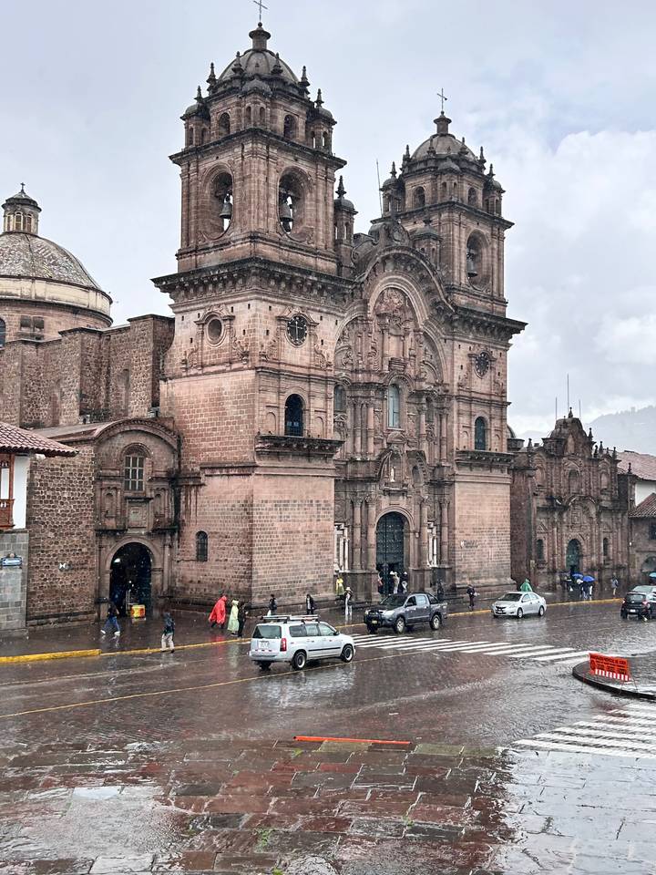 Majestic stone cathedral with baroque façade towering over a rainy city street where pedestrians in raincoats walk by.