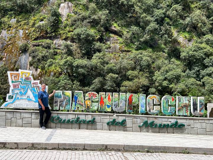 Visitor standing beside the colourful 'MachuPicchu Ciudad del Mundo' sign surrounded by green cliffs.