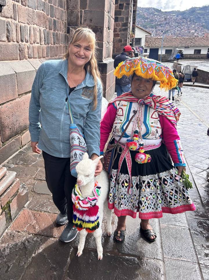 Tourist posing with a traditionally dressed local woman holding a baby alpaca on a cobblestone street.