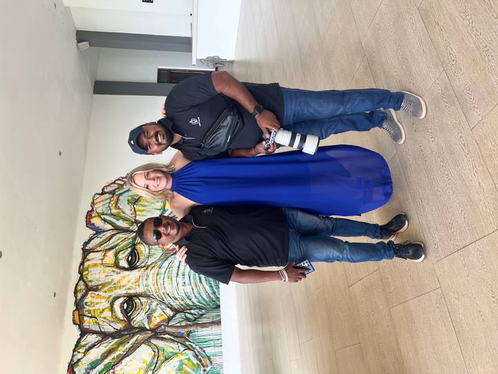 Three people pose indoors beneath a colourful elephant mural, one holding a large camera lens.