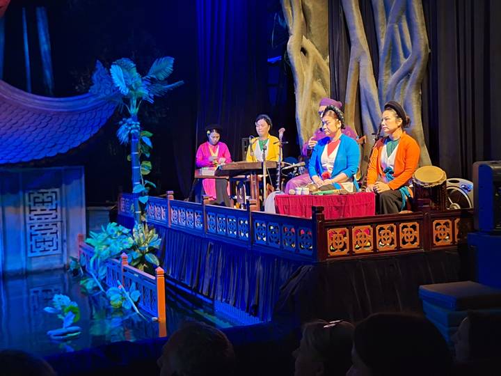 Traditional Vietnamese musicians in colourful attire performing on an indoor stage with instruments.