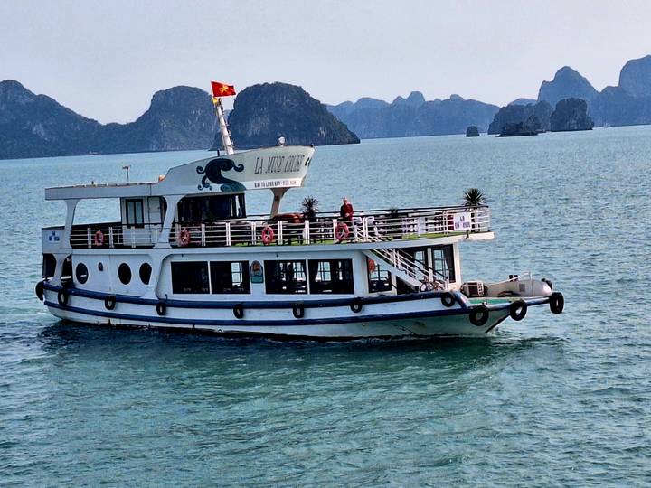 A white sightseeing boat cruising on calm green water with dramatic karst islands in the background and a Vietnamese flag flying.