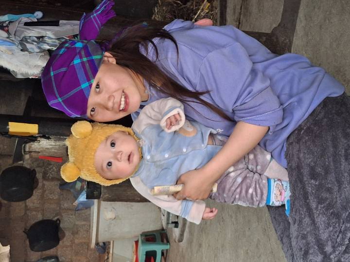 A smiling young woman wearing a colourful headscarf holds a baby in a rustic indoor setting.