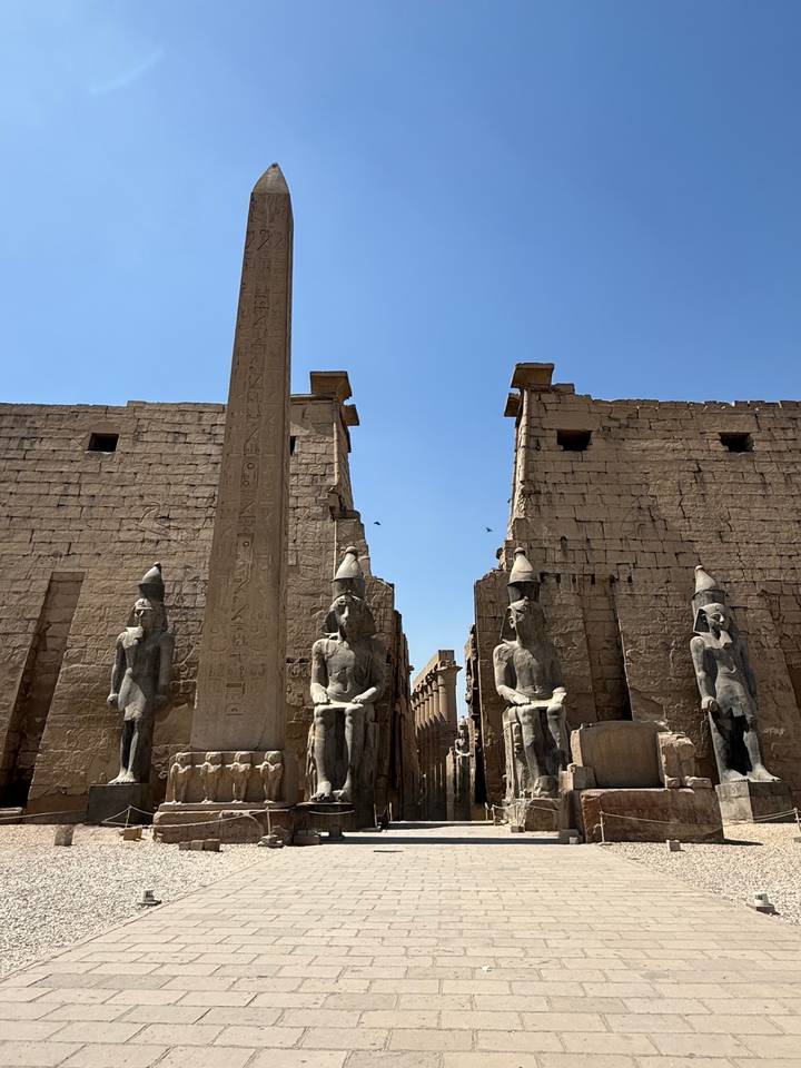 Massive sandstone pylons, statues and a central obelisk stand against a cloudless sky at Luxor Temple.