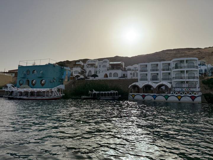 Colourful Nubian guesthouses along the Nile shore glow in the low sun with calm water in the foreground.