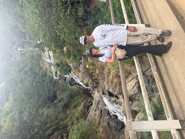 Couple posing on a bridge with a multi-tiered waterfall and lush forested hillside behind them.