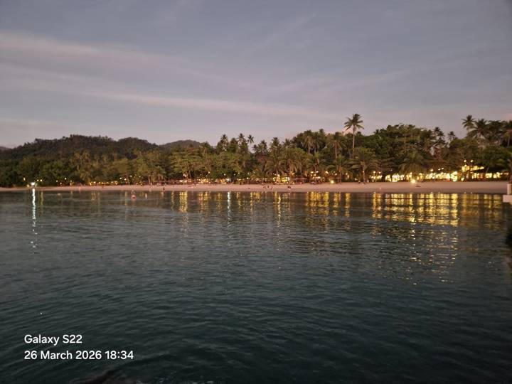 Twilight view from the water of a palm-lined beach illuminated by warm resort lights reflecting on the sea.